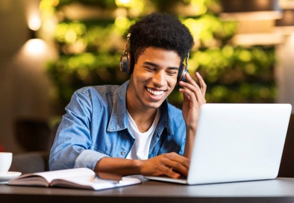 Cheerful black young guy with headset looking at laptop, having fun while studying, cafe interior